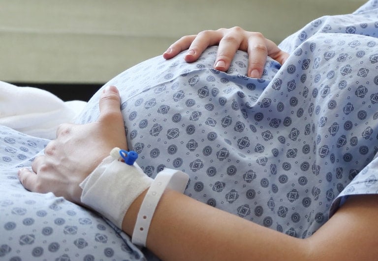 A person in a hospital gown is lying down, holding their pregnant belly with both hands, possibly preparing for a VBAC. An identification bracelet and an IV port are visible on their left wrist. The background is blurred, indicating a medical environment.