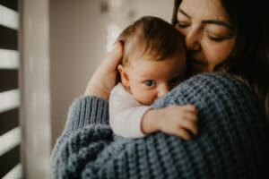 A mother holding her son, Max, close to her chest gently rests her cheek against his head. The baby is wrapped in a soft, gray sweater, looking directly at the camera. The background is softly blurred, emphasizing the tender moment between them.