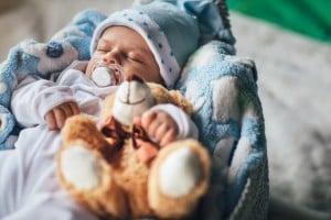 A baby boy wearing a white outfit and a blue hat with star patterns sleeps in a blue and white blanket. The baby has a pacifier in their mouth and is holding a brown stuffed bear, bringing joy to anyone who've longed to conceive a boy.