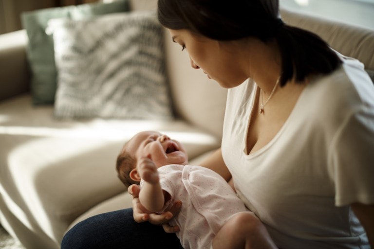 A woman with dark hair is sitting on a couch and holding a baby who is crying, possibly due to baby reflux. The baby is dressed in a white onesie. In the background, there is a beige couch with two pillows. The setting appears to be a living room.