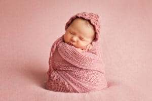 A newborn baby girl, one of those with beautiful y girl names, is swaddled in a pink knitted blanket and wearing a matching pink knitted hat. The baby is peacefully sleeping on a pink background.