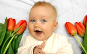 A baby with light skin and blond hair is lying on a light-colored blanket, smiling with wide eyes. The baby is dressed in a white outfit and is surrounded by several red tulips on either side, reminiscent of beautiful flower names for girls like Lily or Rose.