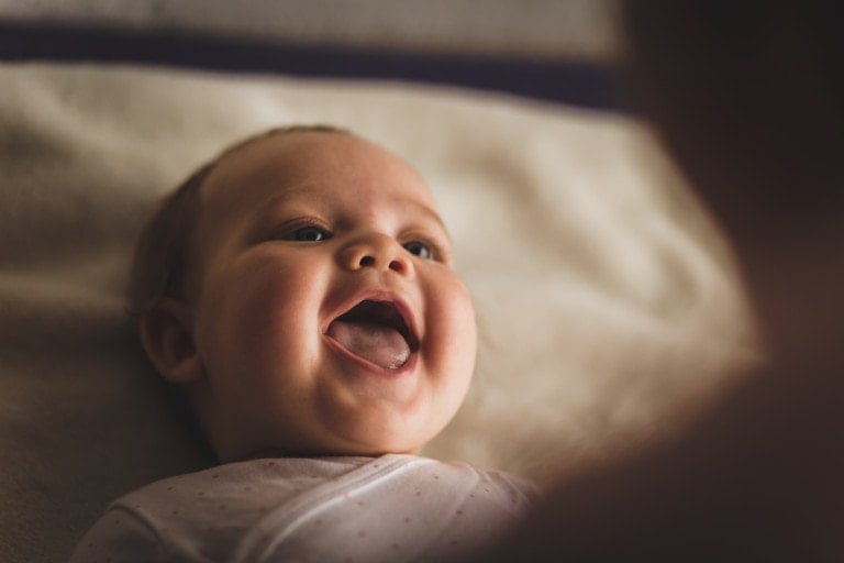 A close-up image of a smiling baby lying down. The baby's mouth is open in a joyful laugh, showing visible gums, and the baby is looking upwards. The background is soft and blurred, implying a cozy and intimate setting. The baby's expression conveys happiness and contentment.