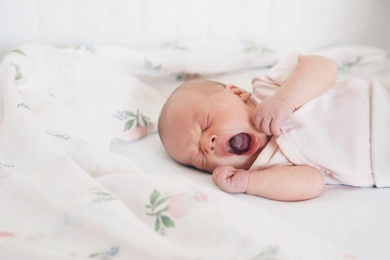 A newborn baby lies on a bed, wrapped in a floral print blanket, and yawning. The baby is wearing a light-colored onesie and has one arm bent while the other rests on its chest. The background is softly focused, featuring more of the floral print—a serene moment untroubled by any thrush symptoms.