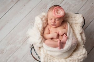 A newborn baby, swaddled in a white blanket, is sleeping peacefully in a cushioned basket. The baby wears a pink headband with a flower. The scene is set on a white wooden floor—a perfect setting to welcome this little one with charming C names for girls like Clara or Chloe.