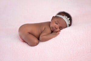 A newborn baby sleeps on their side atop a pink, fluffy blanket. The baby wears a silver headband with a decorative design, and their right hand is tucked under their head. You can almost imagine her parents contemplating r girl names as they watch her sleep peacefully. The background is smooth and matches the blanket.