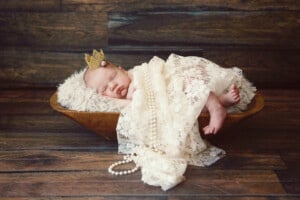 A newborn baby is sleeping in a wooden bowl lined with a fuzzy blanket. The baby, resembling a tiny Greek goddess, is wearing a small crown and wrapped in lace cloth with a strand of pearls draped over them. The background features a rustic, wooden texture.