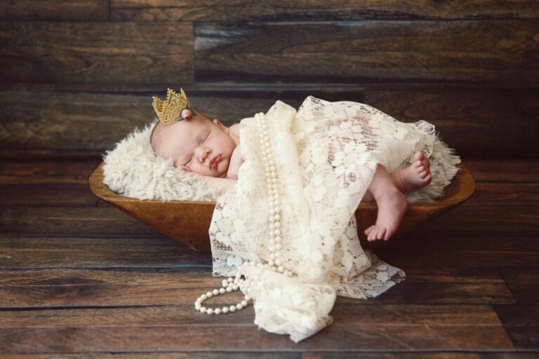 A newborn baby is sleeping in a wooden bowl lined with a fuzzy blanket. The baby, resembling a tiny Greek goddess, is wearing a small crown and wrapped in lace cloth with a strand of pearls draped over them. The background features a rustic, wooden texture.