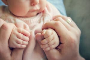 Close-up of an infant in a pink outfit, grasping an adults hands. The adult wears a ring on one finger. The focus is on the hands, with soft lighting creating a warm, gentle feel to the image.