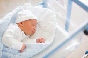 A newborn baby is sleeping on a soft white blanket in a hospital bassinet. The baby is dressed in a white outfit and a white hat adorned with small stars, perfect as one of those cherished coming home outfits. The bassinet has a transparent casing and blue trim.