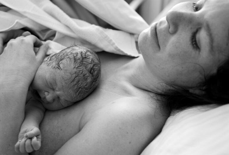 A black-and-white image of a newborn baby resting on their mother's chest after a safe birth. The mother lies in bed, looking up with a calm expression while holding the baby close under a blanket. The baby appears to be crying or making a facial expression.