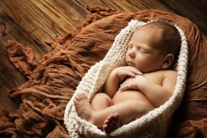 A sleeping baby is swaddled in a knitted cream-colored wrap, lying on a textured brown blanket. The background consists of wooden planks, creating a warm, rustic setting. With hands near its face and legs gently curled, this serene scene is perfect for celebrating gender-neutral names.