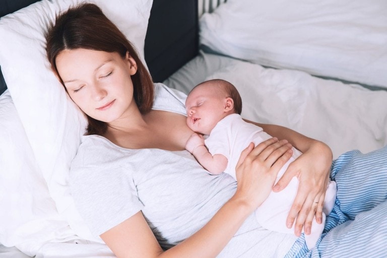 A woman is lying-in on a bed with her eyes closed, resting on a white pillow. She holds a sleeping baby on her chest, supporting the baby with one hand. They are both wearing light-colored clothes, and the background shows white bedding.