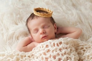 A sleeping baby with dark hair lies on a soft, fluffy white blanket. Wrapped in a knitted cream-colored blanket and wearing a small, gold-colored crown, the baby evokes timeless charm. With arms bent and hands resting near its ears, this little one seems destined for royal names.