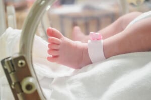 A newborn baby's foot with a pink hospital wristband displaying my baby's name rests inside an incubator. The incubator has a transparent plastic panel and visible metal hinges. The baby's foot is wrapped with a white blanket.