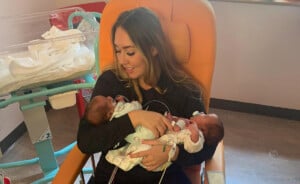 A woman is sitting in an orange chair holding two premature newborn babies, one in each arm. She is looking down at them while smiling. There is a hospital bassinet with white linens and medical equipment in the background.