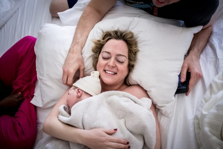 A woman lies in bed, smiling with her eyes closed, cradling a newborn wrapped in a blanket against her chest. A person on each side of the bed touches her shoulder and head gently. This scene captures a tender moment post-childbirth, marking the beginning of her birth story as she became a mother.