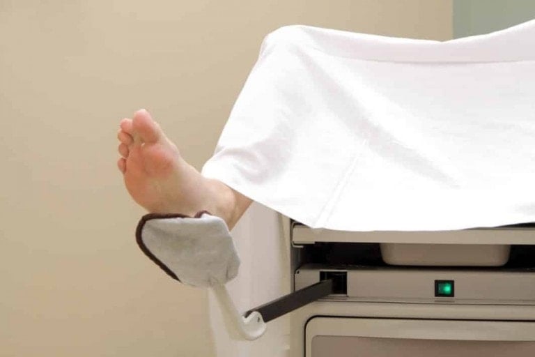 A person's bare foot protrudes from under a white sheet while resting in a cushioned footrest attached to a medical examination table in a clinical setting, poised for a cervical check.