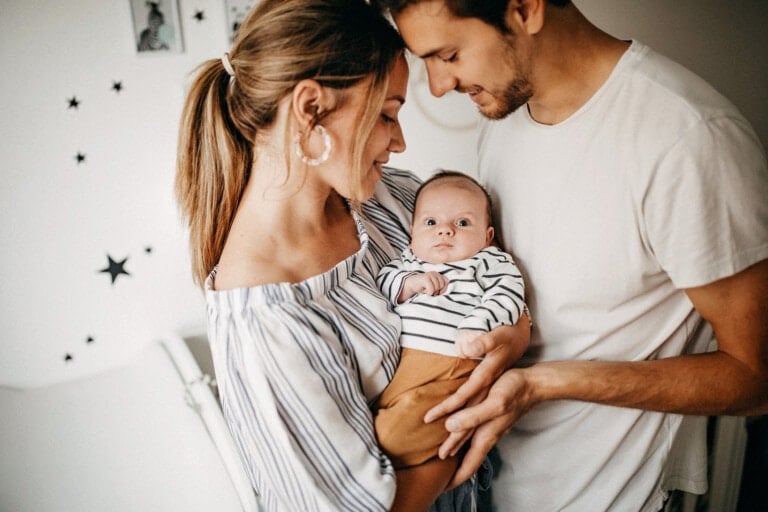 A man and woman stand close together, looking down lovingly at a baby they are holding. The woman, wearing a striped off-shoulder top, and the man in a white T-shirt embody relationship advice with their nurturing bond. The baby is adorably dressed in a matching striped outfit.