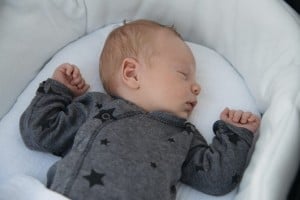 A sleeping baby lies in a white bassinette, wearing a gray onesie with black star patterns. The baby has light-colored hair and rests with both arms bent, each hand placed near their head, peacefully sleeping through the night. The bedding is simple and white.
