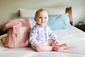 A baby with a short haircut and wearing a plaid shirt sits on a bed next to a pink diaper bag. The bed, adorned with white and blue bedding, complements the brown headboard in the background. This little one looks like he could have one of those preppy boy names that are all the rage right now.
