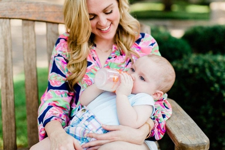 A woman with long blonde hair, wearing a bright, colorful blouse, sits on a wooden bench outdoors. She is holding a baby who is drinking from a PopYum bottle. The baby is wearing a light blue onesie and checkered shorts. Both appear to be relaxed and content.