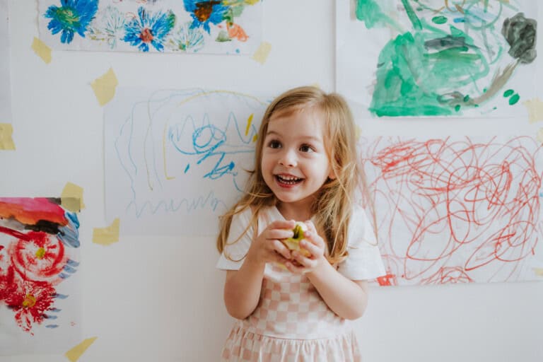 A young child with long hair, wearing a checkered dress, stands in front of a wall adorned with various colorful children artwork taped to it. The child is holding an object and smiling joyfully.
