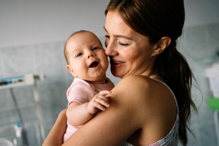 All I do as a new mom is captured in this tender moment: a woman holding an infant who is smiling. She looks at her baby affectionately, and the baby, dressed in a light pink onesie, beams back. The background features a tiled wall and some medical equipment.
