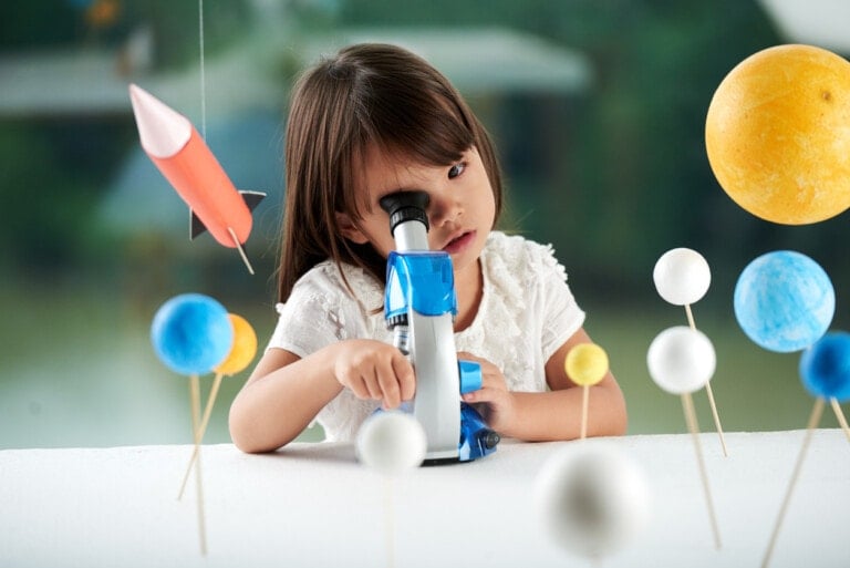 A young child in a white shirt looks through a microscope. Around them are various models of planets and a rocket, indicating a space-themed setting. The background is blurred, focusing attention on the child and the objects in the foreground, showcasing some of the best STEM toys for budding scientists.