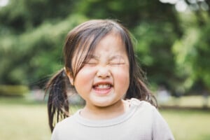 A young girl with pigtails, possibly named Willow or Wendy, is standing outdoors with her eyes closed and mouth open in a cheerful expression. The background is blurred, showing some green foliage. The child is wearing a light-colored shirt.