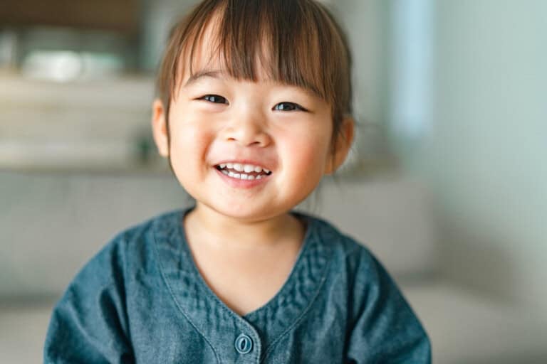 A young child with straight brown hair, bangs, and a blue shirt smiles while sitting indoors. The softly blurred background highlights a neutral-colored couch and wall—perfect for capturing special 2-year-old milestones.