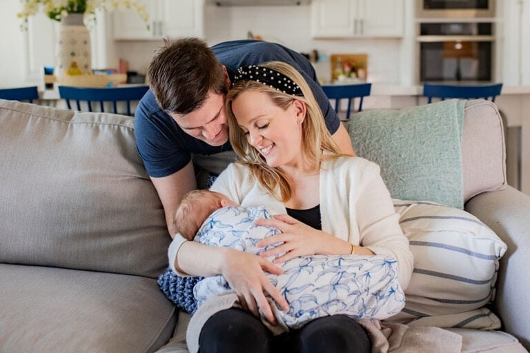 A woman is sitting on a couch, cradling a baby wrapped in a blue and white blanket. A man is leaning over her shoulder, smiling at the baby. The scene takes place in a bright, modern living room designed to help new moms during postpartum recovery.