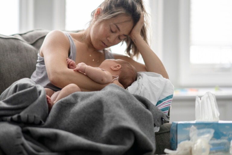 A tired woman experiencing mom burnout sits on a couch, head resting on her hand as she breastfeeds her baby. Covered with a grey blanket, there's a box of tissues by her side. The background reveals a bright room with windows.