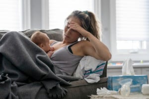 A woman with a ponytail is sitting on a couch, covered with a blanket, holding a baby and appearing tired. Resting her head on one hand, she looks like she's trying to figure out how to stay sane when the baby won't nap. A box of tissues and a breast pump sit on the table next to her in the naturally lit room.