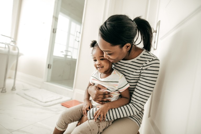 A woman with a ponytail, wearing a striped shirt, holds and kisses a smiling toddler on the cheek. They are sitting on the floor in a brightly lit bathroom with a glass shower door and white decor, celebrating that the little one was potty trained in one day.