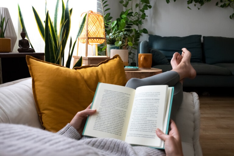 A person is lying on a couch holding an open book. The room has a cozy atmosphere with a mix of potted plants, a wooden table, a yellow cushion, and a small lamp. The relaxed setting feels perfect for spending Mother's Day, with the couch facing a coffee table with a mug on it and another couch visible in the background.