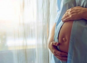 Close-up of a pregnant woman's belly. She is wearing a light blue delivery gown and cradling her belly with both hands. She stands near a sheer curtain, with soft natural light coming through. The setting appears calm and serene.