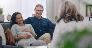A pregnant woman and a man sit closely together on a couch, smiling. The woman has her hand on her belly. They are in a well-lit room, speaking to a person whose back is to the camera. The setting appears to be a relaxed office space, perhaps for a relationship checkup before the baby arrives.