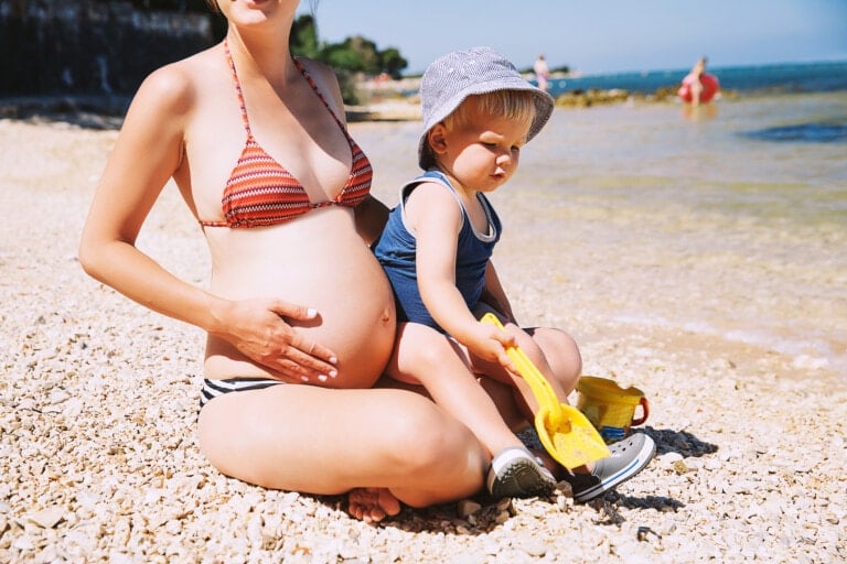 A pregnant woman sits on a pebbled beach wearing a striped bikini, enjoying her babymoon. A young child in a hat and swimwear sits on her lap, playing with a yellow toy shovel and a blue bucket. Other people are seen in the background near the water.