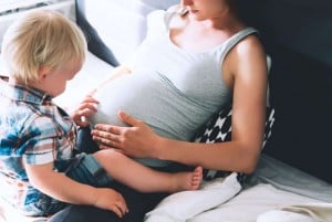 A pregnant woman in a gray tank top is sitting on a bed, with a young child in a plaid shirt on her lap. The child touches her belly, curious about how pregnancy changes as you get older. They both appear to be in a relaxed, home setting.