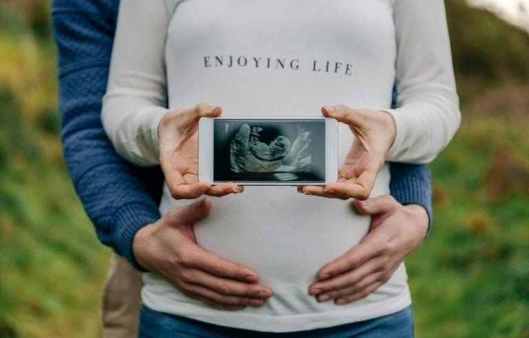 A pregnant person wearing a white shirt with the text "ENJOYING LIFE" holds an ultrasound image. Another person's hands gently rest on the pregnant person's belly from behind, making this a beautiful pregnancy announcement in an outdoor, grassy area.