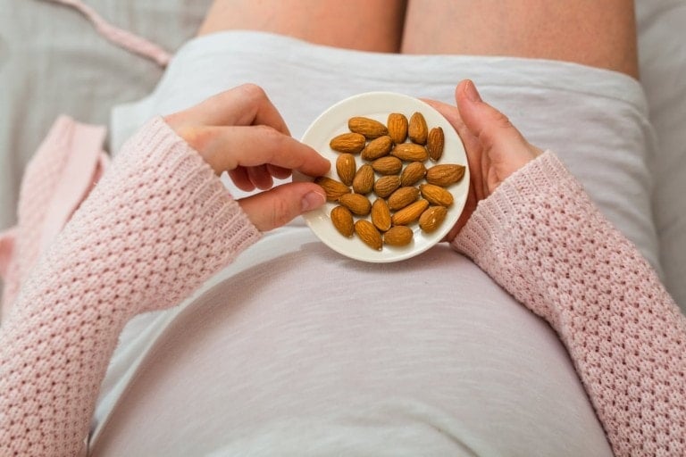 A person wearing a pink knit sweater is seated, holding a small white bowl of almonds with both hands. The bowl is held over their lap, and one almond is being picked up with the right hand. The background appears to be a bed, suggesting a cozy setting for enjoying pregnancy snacks.