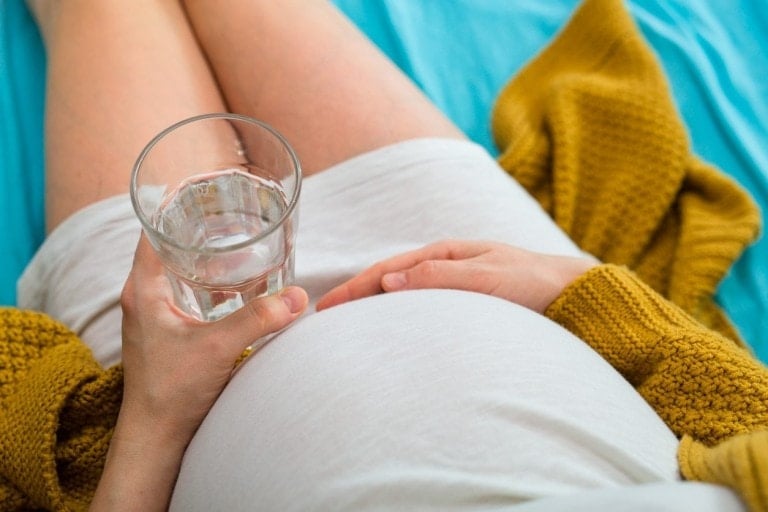 A pregnant person wearing a white dress and a yellow cardigan holds a glass of water while resting on a blue sheet, mindful of avoiding dehydration in pregnancy. Their other hand is placed gently on their belly.
