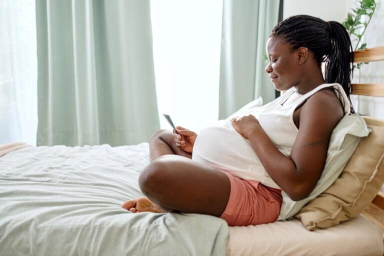 A pregnant woman is sitting comfortably on a bed, leaning against pillows. She is looking at a smartphone and smiling. The room has light-colored curtains and bedding. She is wearing a white top and pink shorts.