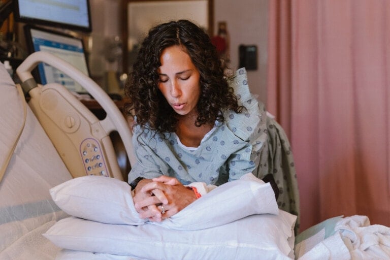 A woman with curly hair is leaning over a hospital bed, looking down. She is dressed in a hospital gown and appears to be in a hospital room with medical equipment visible in the background. She’s holding onto a pillow positioned on the bed, possibly practicing labor breathing techniques.