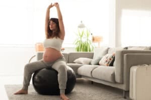 A pregnant woman sits on a black exercise ball, arms stretched overhead, as she prepares her body for labor and birth. She wears a white top and gray sweatpants. The room features a gray sofa, a plant, and soft lighting.