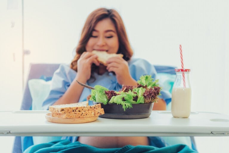A person in hospital attire sits on a bed, eating a sandwich. In the foreground, there is a tray with sliced bread, a bowl of salad, and a bottle of milk with a red-striped straw. The setting appears to be a hospital room, prompting the question: can you eat during labor?