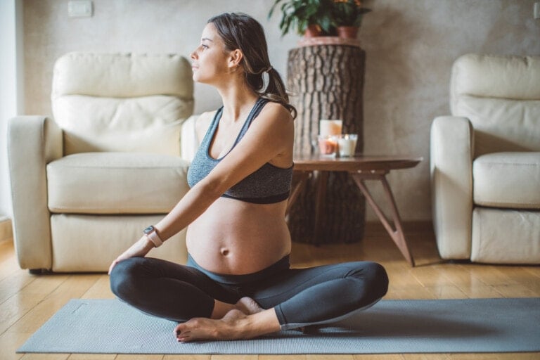 A pregnant woman is seated cross-legged on a yoga mat indoors, performing a seated twist to make childbirth easier. She is wearing a sports bra and leggings, and looking out of the window. Two beige armchairs and a wooden table with candles and plants are visible in the background.