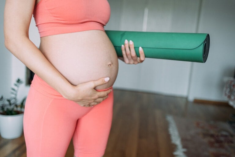 A pregnant person in a pink athletic outfit holds a green yoga mat with one hand while touching their belly with the other, embodying the mindful practice of pilates during pregnancy. The scene is indoors, featuring a wooden floor and a blurred background with a plant.