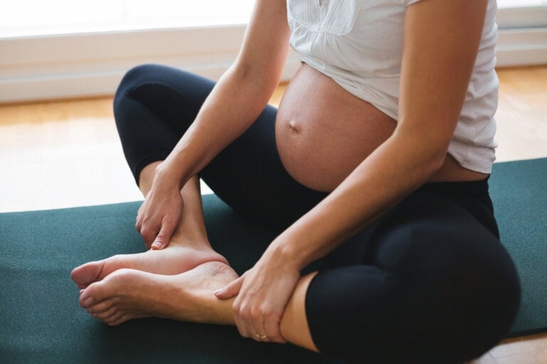 A pregnant person sits cross-legged on a yoga mat indoors, wearing a white sleeveless top and black leggings. One hand rests on their shin, and the other hand touches their foot. The background is softly lit by natural light through a window.